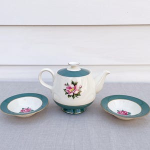 May include: A vintage tea set comprising a white teapot with a teal lid and base, and two matching saucers. Each item is decorated with a pink floral pattern. The set is displayed on a light grey surface.