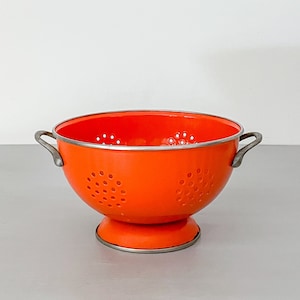 May include: Orange enamel colander with silver handles and a silver rim. The colander has a round base and is made of metal.
