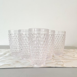 Könnte beinhalten: Vier klare Glasbecher mit einer strukturierten, geblähten Oberfläche. Die Gläser sind in einer Reihe auf einer weißen und goldfarbenen, gemusterten Oberfläche angeordnet.