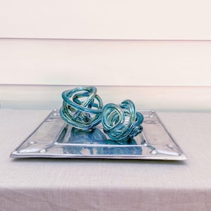 May include: Two decorative glass knot sculptures in shades of teal and blue rest on a square, silver-toned metal tray. The sculptures have a woven appearance. The tray has a riveted edge and is placed on a light gray surface.