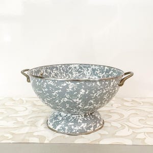 May include: A gray and white enamelware bowl with a pedestal base and two brass handles. The bowl has a speckled pattern and a metal rim. This vintage-style bowl is suitable for kitchen or home decor.