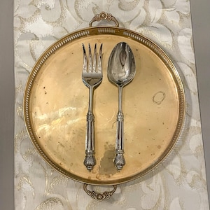 Könnte beinhalten: Ein goldfarbenes Serviertablett aus Metall mit einer Gabel und einem Löffel. Die Utensilien haben verzierte silberne Griffe mit einem burgunderfarbenen Streifen. Das Tablett liegt auf einer weiß-gold gemusterten Tischdecke.