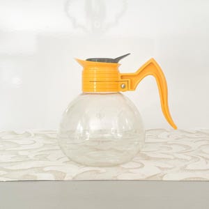 Może przedstawiać: Przezroczysty szklany dzbanek do kawy o zaokrąglonym korpusie i jasnożółtym uchwycie i pokrywce. Uchwyt ma zakrzywioną, otwartą konstrukcję. Pokrywka jest czarna. Karafka stoi na wzorzystej powierzchni.