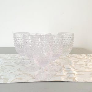 Könnte beinhalten: Satz von fünf klaren Glaskelchen mit einer strukturierten, geblähten Oberfläche. Die Kelche sind auf einem weißen Tischläufer mit Blumenmuster angeordnet.