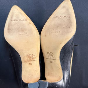 May include: A pair of black leather Manolo Blahnik heels, size 38, with pointed toes and small heels. The soles are beige with the brand name and "Made in Italy" printed on them. The heels have a leather upper and leather sole.
