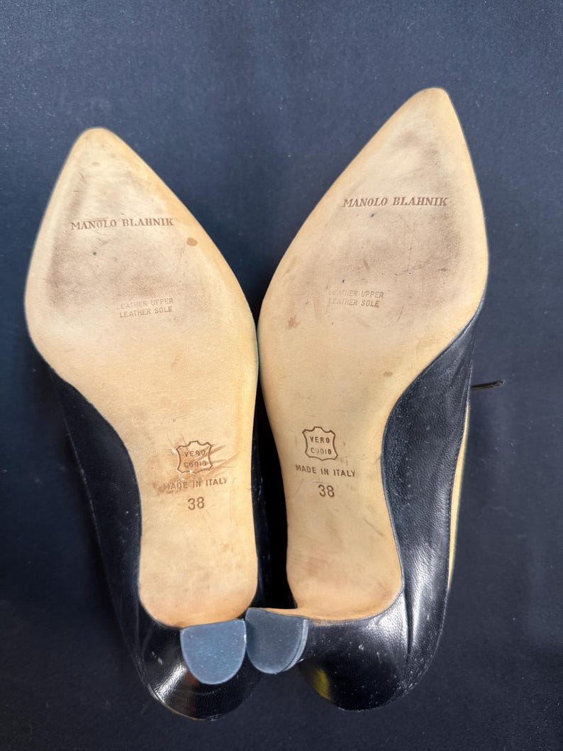 May include: A pair of black leather Manolo Blahnik heels, size 38, with pointed toes and small heels. The soles are beige with the brand name and "Made in Italy" printed on them. The heels have a leather upper and leather sole.