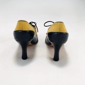 May include: A pair of two-tone, heeled oxfords. The shoes feature a mustard yellow suede upper and black leather detailing on the toe and heel. The heels are black and slender. The shoes are laced with black laces.
