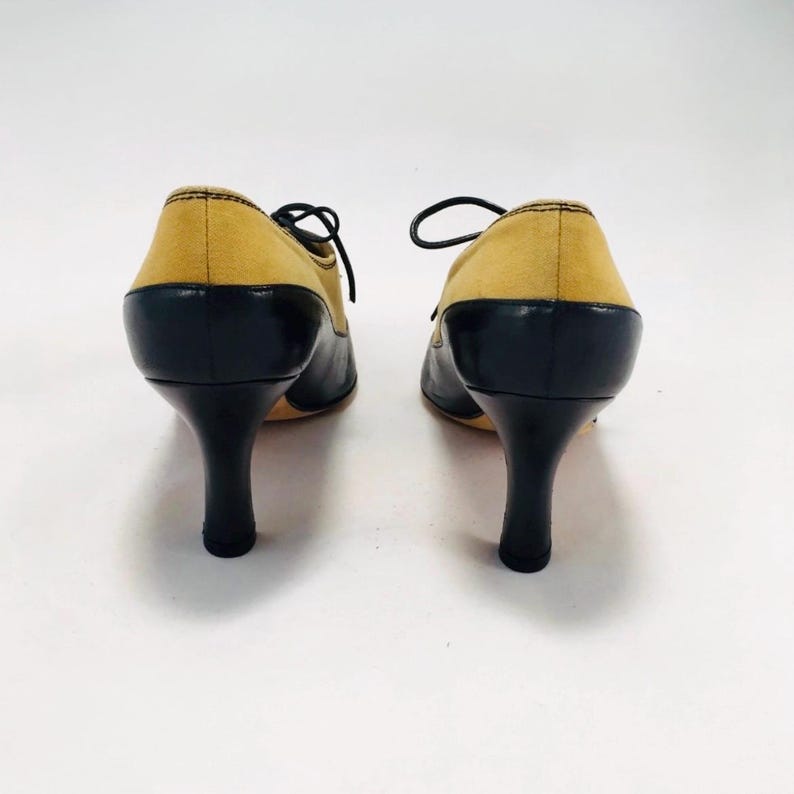 May include: A pair of two-tone, heeled oxfords. The shoes feature a mustard yellow suede upper and black leather detailing on the toe and heel. The heels are black and slender. The shoes are laced with black laces.