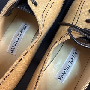 May include: Close-up of two tan canvas shoes with black stitching and black leather accents. The shoes have black laces and the brand name "MANOLO BLAHNIK" is printed on a white label inside each shoe.