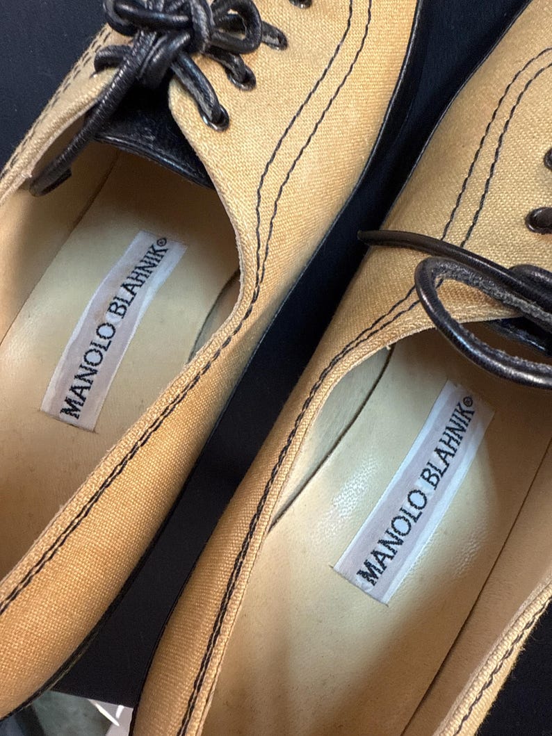 May include: Close-up of two tan canvas shoes with black stitching and black leather accents. The shoes have black laces and the brand name "MANOLO BLAHNIK" is printed on a white label inside each shoe.