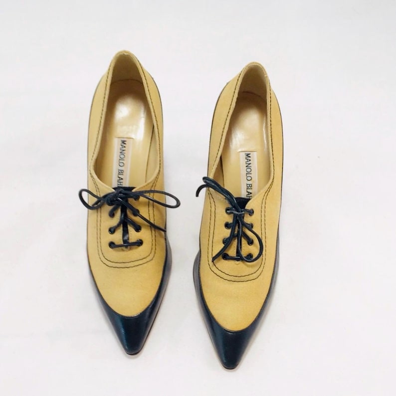 May include: A pair of tan and black leather heeled oxfords. The shoes have a pointed toe and black laces. The brand name "MANOLO BLAHNIK" is printed on the insole. The shoes are set against a white background.
