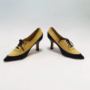 May include: A pair of two-tone, pointed-toe pumps. The shoes feature a tan suede upper with black patent leather accents on the toe and heel. Black laces and a stiletto heel complete the design. The shoes are set against a white background.