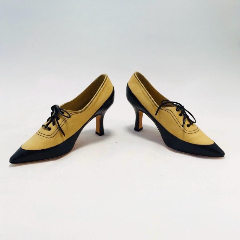 May include: A pair of two-tone, pointed-toe pumps. The shoes feature a tan suede upper with black patent leather accents on the toe and heel. Black laces and a stiletto heel complete the design. The shoes are set against a white background.