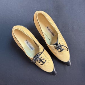 May include: A pair of tan suede Manolo Blahnik shoes with black leather toe caps and black laces. The shoes have a pointed toe design and the brand name is visible inside the shoe.