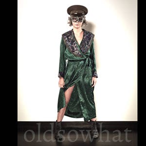 May include: A person wearing a dark green velvet wrap dress with a floral embroidered collar and a matching belt. The person is also wearing a brown hat with goggles and black ankle boots. The text "oldsowhat" is at the bottom.