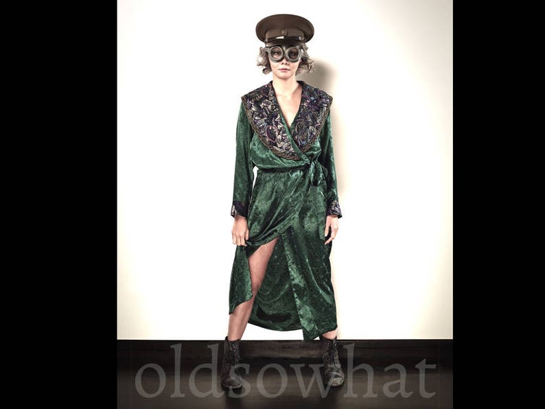 May include: A person wearing a dark green velvet wrap dress with a floral embroidered collar and a matching belt. The person is also wearing a brown hat with goggles and black ankle boots. The text "oldsowhat" is at the bottom.