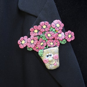 Puede incluir: Un broche de flores rosa y amarillo con una cara sonriente en la maceta. El broche está hecho de tela y está sujeto a una chaqueta negra.