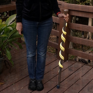 Op de afbeelding: Een houten wandelstok met een geel en zwart spiraalontwerp. De stok wordt vastgehouden door een persoon die een blauwe spijkerbroek en een zwart jack draagt.