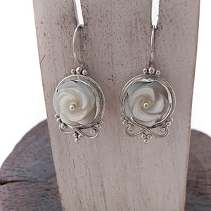 Peut inclure: Une paire de boucles d'oreilles en argent avec un motif de rose en nacre blanche. Les boucles d'oreilles sont suspendues à un support en bois.