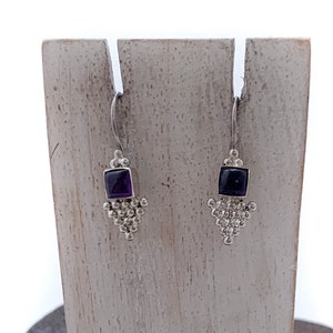 May include: A pair of silver dangle earrings with a small square amethyst gemstone in the centre of each earring. The earrings are displayed on a wooden stand.