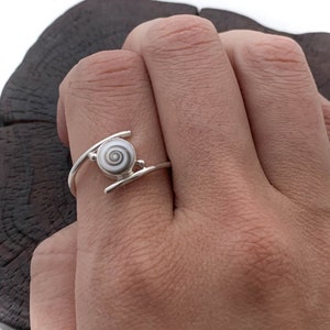 May include: A silver ring featuring a white seashell set in the centre. The ring is worn on a person's finger.