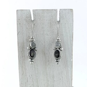 May include: A pair of silver dangle earrings with a dark gray oval gemstone and smaller round clear gemstones. The earrings are displayed on a white wooden stand.