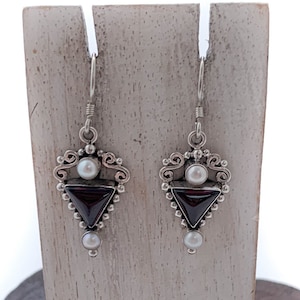 May include: A pair of silver earrings featuring a triangular garnet stone and a pearl accent. The earrings are displayed on a wooden stand.