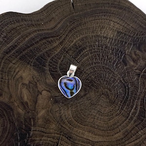 Peut inclure: Un pendentif en argent en forme de cœur avec une incrustation de nacre bleue et verte. Le pendentif est présenté sur une surface en bois foncé.