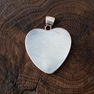 May include: A silver heart-shaped pendant with a smooth, polished surface. The pendant is displayed on a dark brown wooden surface.