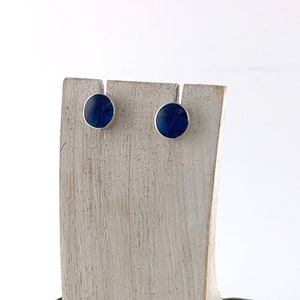 Peut inclure: Une paire de petites boucles d'oreilles rondes bleues avec des montures argentées. Les boucles d'oreilles sont présentées sur un support en bois.