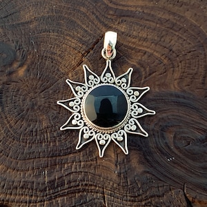 May include: A silver pendant featuring a black onyx stone in the centre, shaped like a sun with intricate details. The pendant is displayed on a wooden surface.