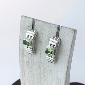 May include: A pair of silver hoop earrings with green gemstone accents. The earrings are displayed on a wooden stand.