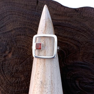 May include: A silver ring with a square red gemstone set in a silver frame. The ring is displayed on a wooden ring holder.