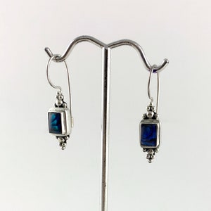 May include: Silver earrings featuring rectangular, blue-green iridescent stones. The earrings are displayed on a silver stand with a clear acrylic base, set on a dark wooden surface. Each earring has a small decorative bead at the bottom.