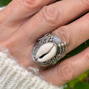May include: A silver ring with a white seashell centre. The ring has detailed patterns around the shell, set against green leaves. The ring is worn on a hand with painted nails.