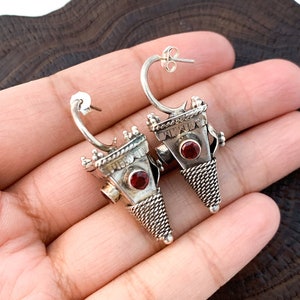 May include: A pair of silver earrings with a red stone in the centre of each earring. The earrings are shaped like a triangle and have a detailed design.