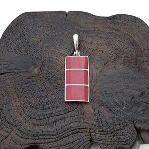 May include: A silver pendant featuring a rectangular red gemstone. The pendant is displayed on a dark wood surface.
