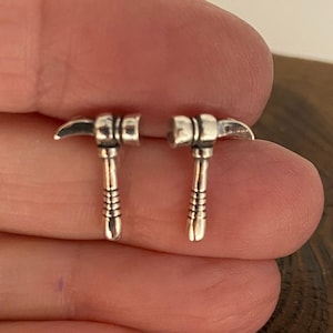 May include: A pair of silver pickaxe stud earrings. The pickaxes feature a detailed design with a silver head and a copper-coloured handle. These small, delicate earrings are suitable for everyday wear.