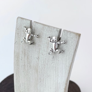 May include: A pair of silver frog earrings displayed on a white wooden stand. The stand is on a dark brown wooden surface.
