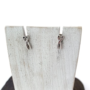 May include: A pair of silver earrings with a simple, intertwined design. The earrings are displayed on a white wooden stand.