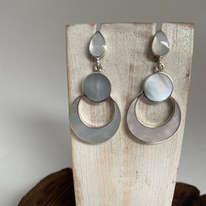 Peut inclure: Une paire de boucles d'oreilles en argent avec une pierre en nacre en forme de larme en haut et deux pierres en nacre rondes plus grandes au centre. Les boucles d'oreilles sont suspendues à un crochet en argent.