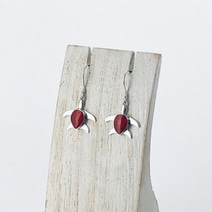 May include: A pair of silver turtle earrings with red gemstone accents displayed on a white wooden stand.
