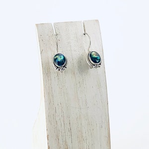 May include: A pair of silver dangle earrings with iridescent blue and green stones. The earrings are displayed on a white wooden stand.