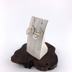 Op de afbeelding: Een paar zilveren oorbellen met een rond ontwerp met kleine witte parels. De oorbellen worden gepresenteerd op een witte houten standaard.