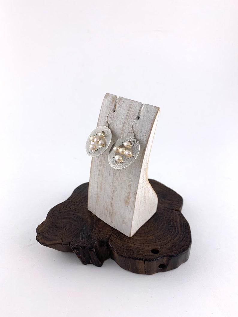 Op de afbeelding: Een paar zilveren oorbellen met een rond ontwerp met kleine witte parels. De oorbellen worden gepresenteerd op een witte houten standaard.