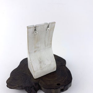 May include: A pair of silver stud earrings with clear stones, displayed on a white wooden earring stand. The stand is on a dark brown wooden base.
