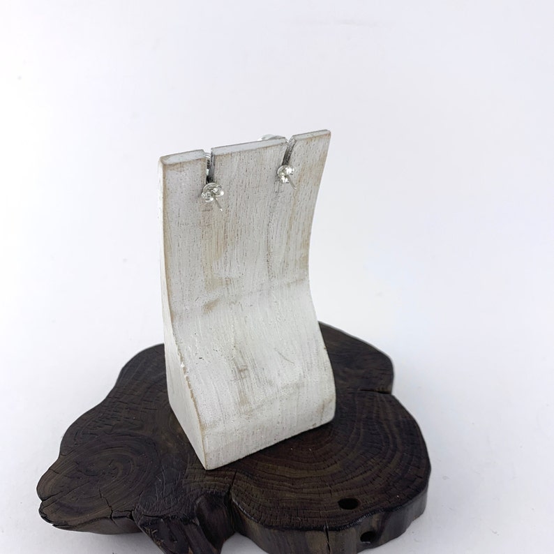 May include: A pair of silver stud earrings with clear stones, displayed on a white wooden earring stand. The stand is on a dark brown wooden base.