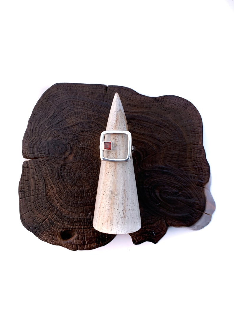 May include: A silver ring with a square red gemstone set in a silver frame. The ring is displayed on a wooden ring holder.