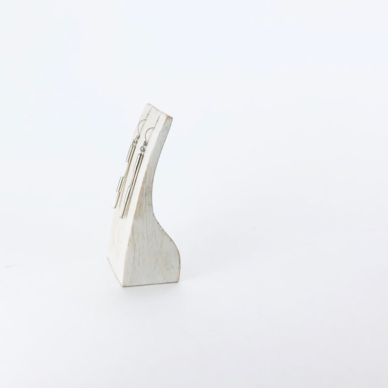 May include: A pair of silver dangle earrings with a cylindrical design displayed on a white wooden stand.