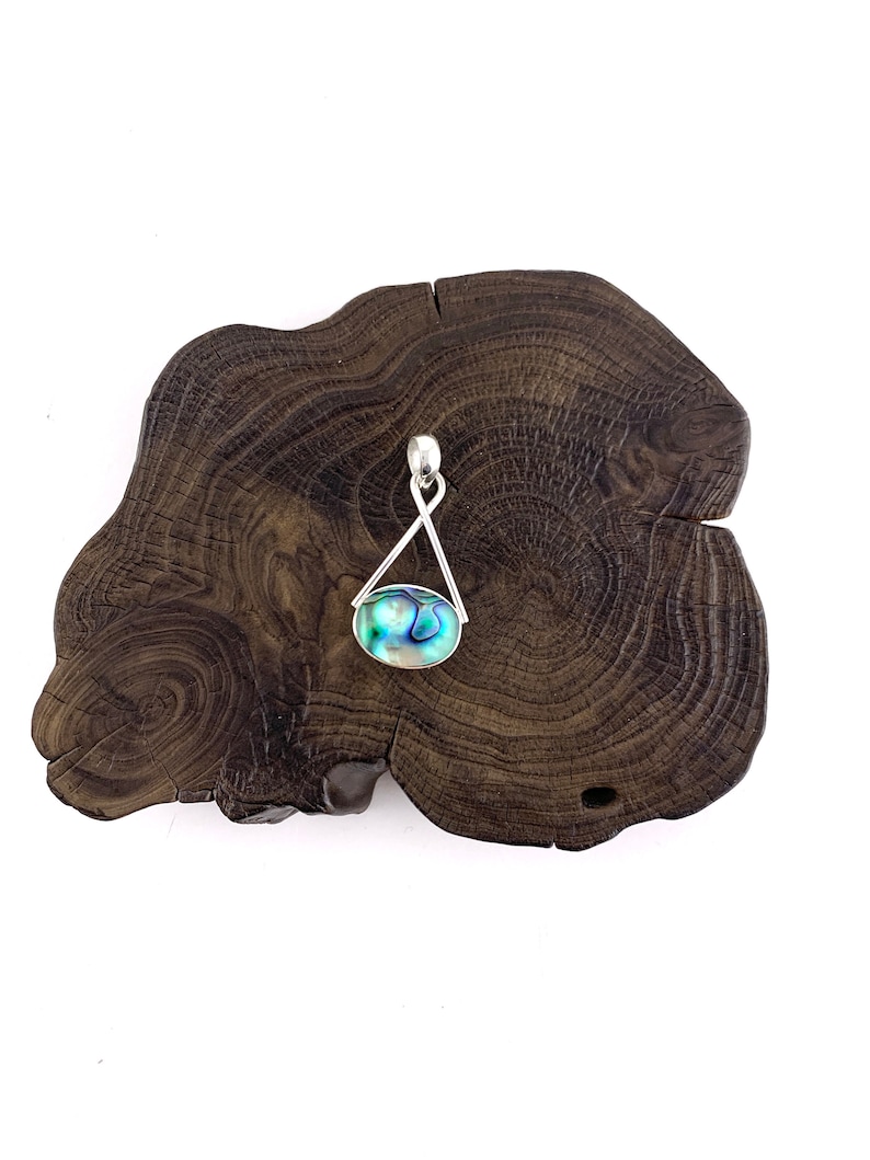 May include: A silver pendant with a blue and green abalone shell, hanging from a silver chain. The pendant is displayed on a dark brown wooden surface.